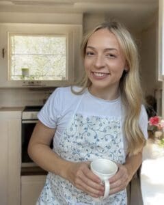Blonde woman holding a cup of tea in front of a kitchen.